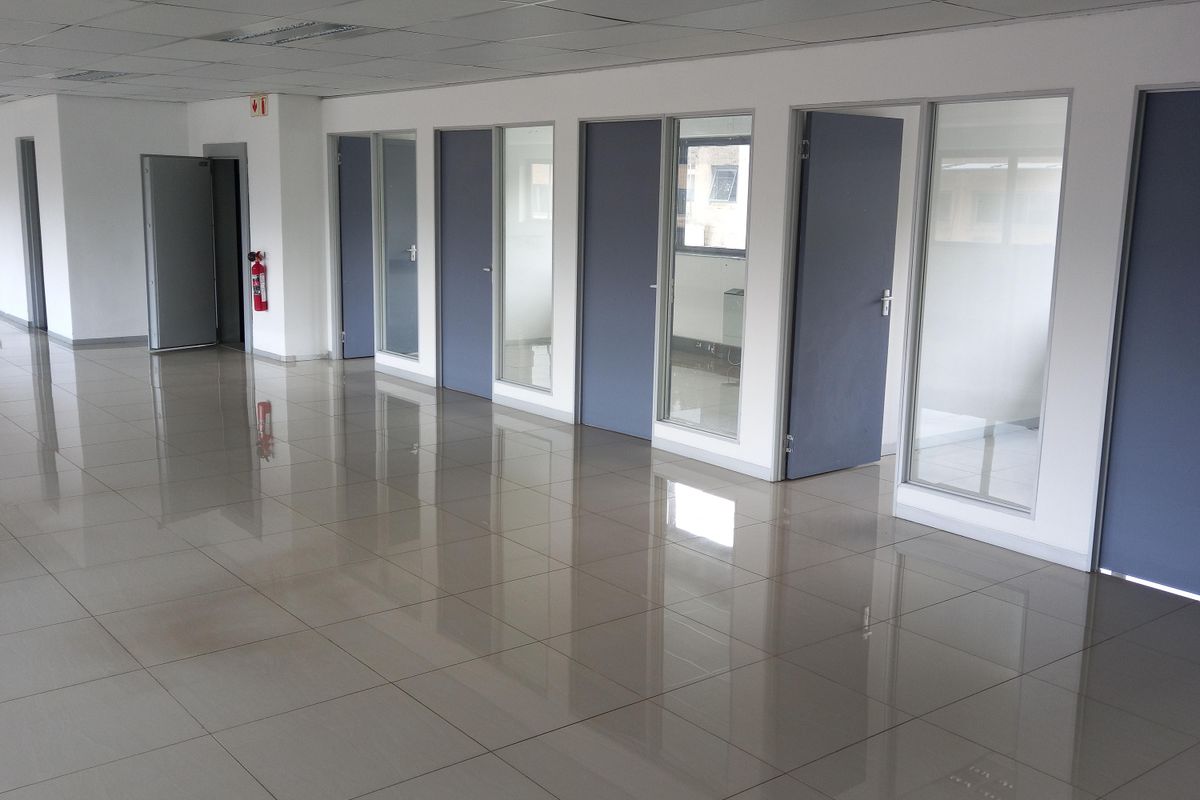 A spacious, empty office area featuring polished tiled flooring, suspended ceiling tiles, and multiple glass-paneled partition doors.