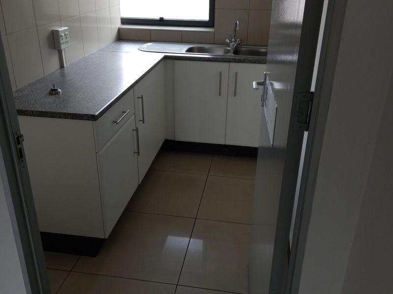 A compact, functional office kitchenette featuring white built-in cabinetry, laminate countertops, a stainless steel sink, and tiled flooring.