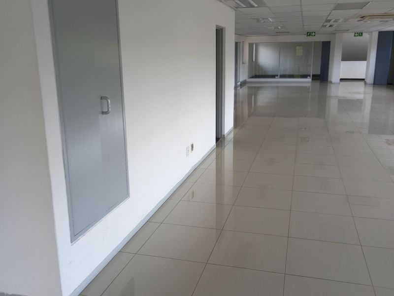 A spacious, empty office hallway featuring polished ceramic tile flooring, a suspended ceiling with recessed lighting, and integrated fire safety equipment.