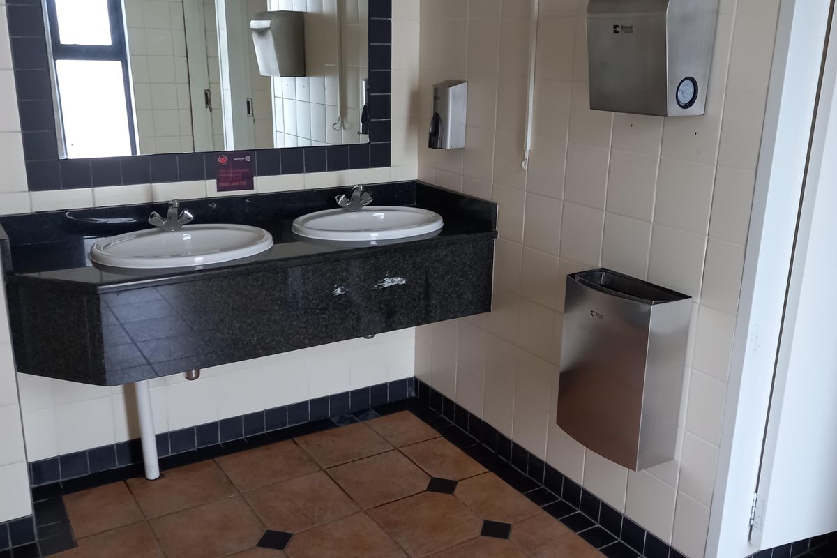 Office restroom featuring a dual-basin vanity with granite countertop, wall-mounted soap and paper towel dispensers, and tiled flooring.
