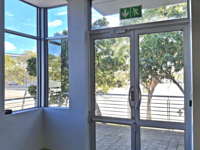 A bright, functional industrial entry area featuring aluminium-framed glass double doors, large windows, and a clear emergency exit sign.