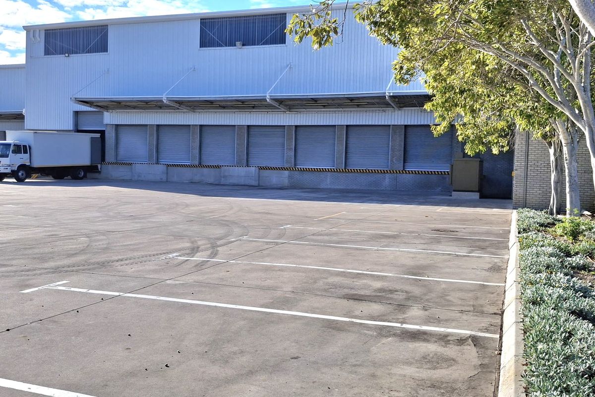 Industrial warehouse exterior featuring multiple roller shutter loading bays, a protective canopy, and a large concrete yard area with marked parking bays.