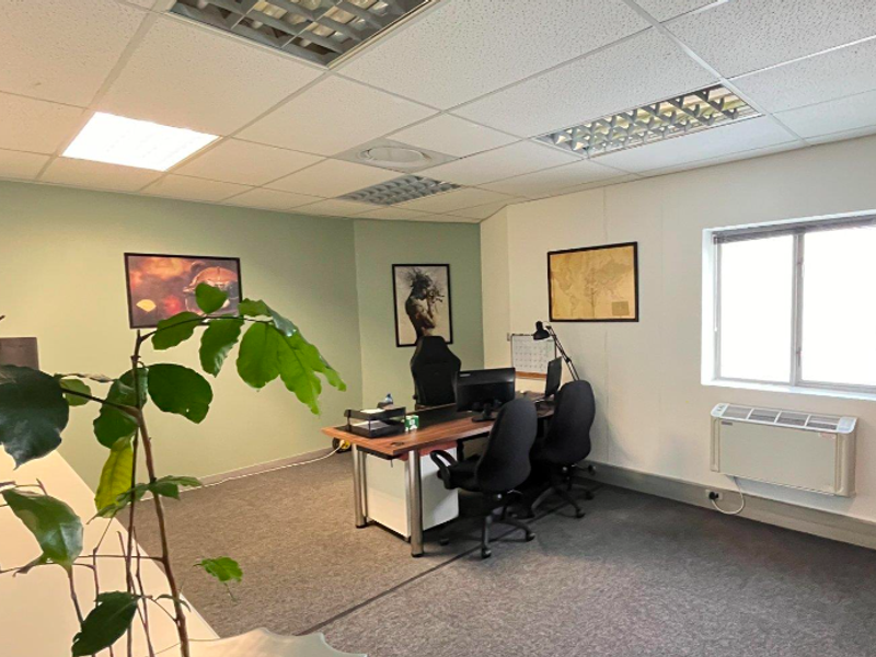 A functional office space featuring carpeted flooring, a suspended ceiling with recessed lighting, and a wall-mounted air conditioning unit.
