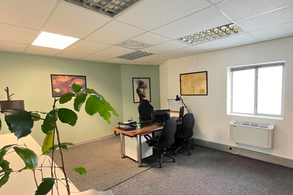 A functional office space featuring carpeted flooring, a suspended ceiling with recessed lighting, and a wall-mounted air conditioning unit.
