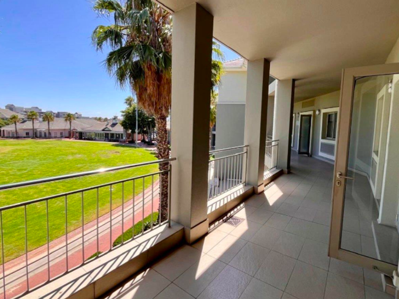 A spacious covered balcony featuring tiled flooring, metal balustrades, and concrete pillars, offering an open view of the surrounding office park grounds.