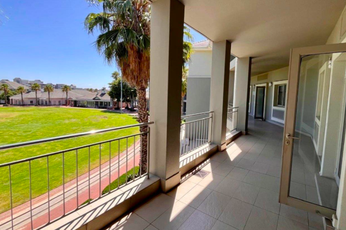 A spacious covered balcony featuring tiled flooring, metal balustrades, and concrete pillars, offering an open view of the surrounding office park grounds.