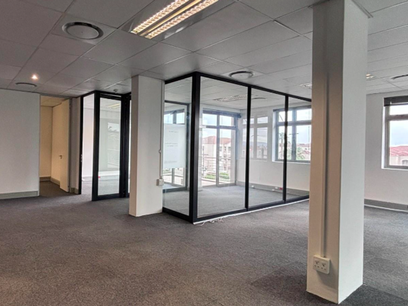 Spacious open-plan office area featuring glass-partitioned meeting rooms, suspended ceilings with recessed lighting, and large windows providing natural light.