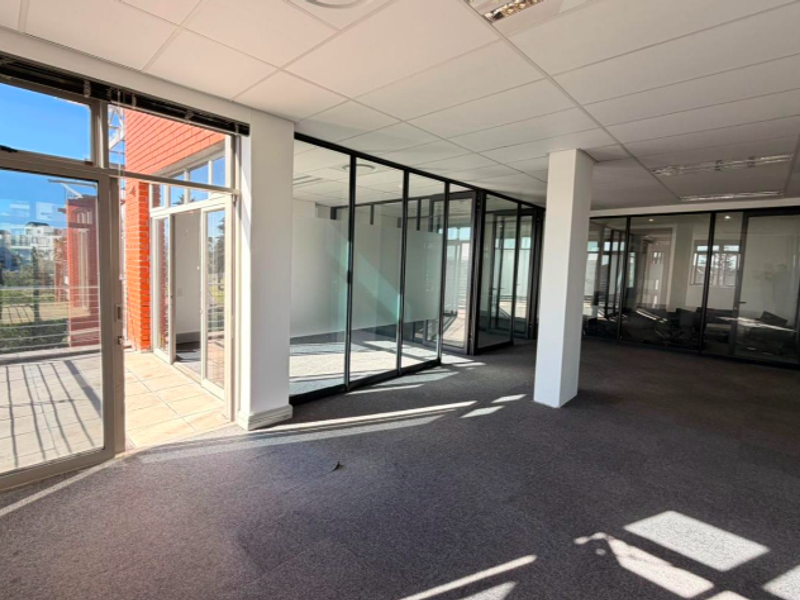 Modern open-plan office space featuring glass-partitioned meeting rooms, carpeted flooring, suspended ceilings, and direct access to an outdoor balcony area.