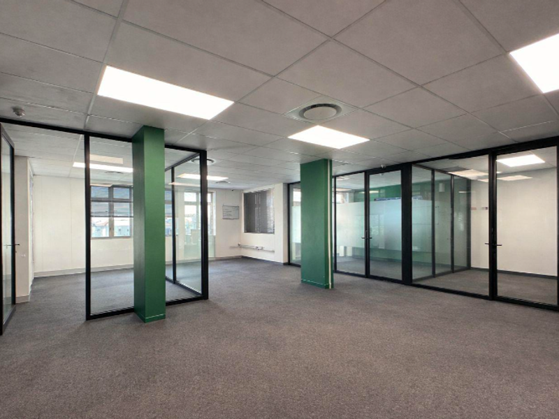 A spacious, modern open-plan office area featuring glass partitions, suspended ceilings with recessed lighting, and neutral carpeting.