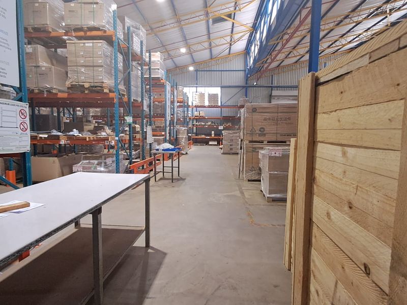 Spacious industrial warehouse featuring high-bay lighting, exposed steel roof trusses, and extensive heavy-duty steel racking systems on concrete flooring.