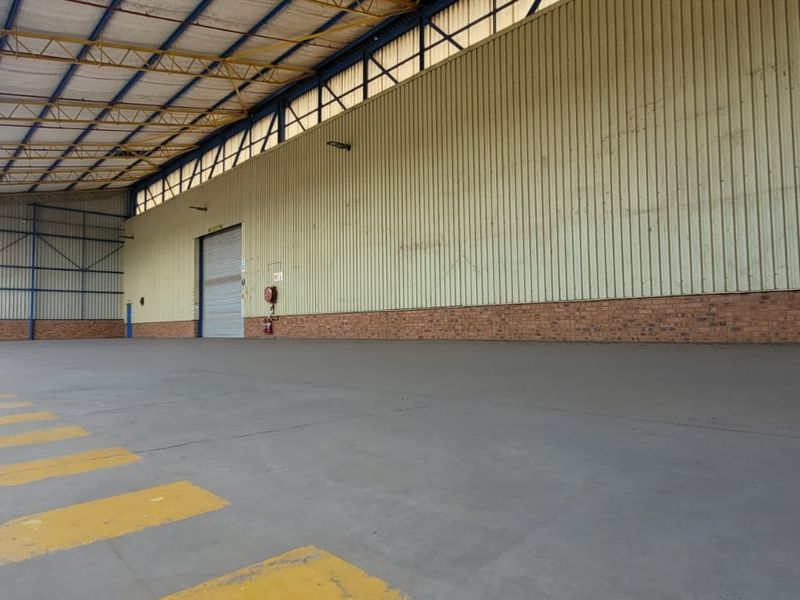 Spacious industrial warehouse unit featuring high ceilings with exposed steel trusses, concrete flooring, and a large roller shutter door for easy access.