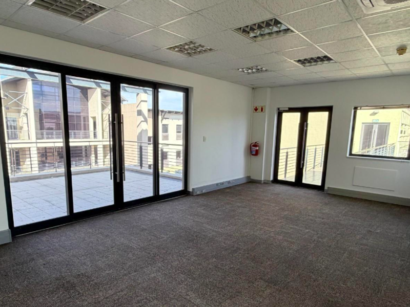 A bright, empty office space featuring carpeted flooring, a suspended ceiling with recessed lighting, and direct access to a private balcony through sliding glass doors.