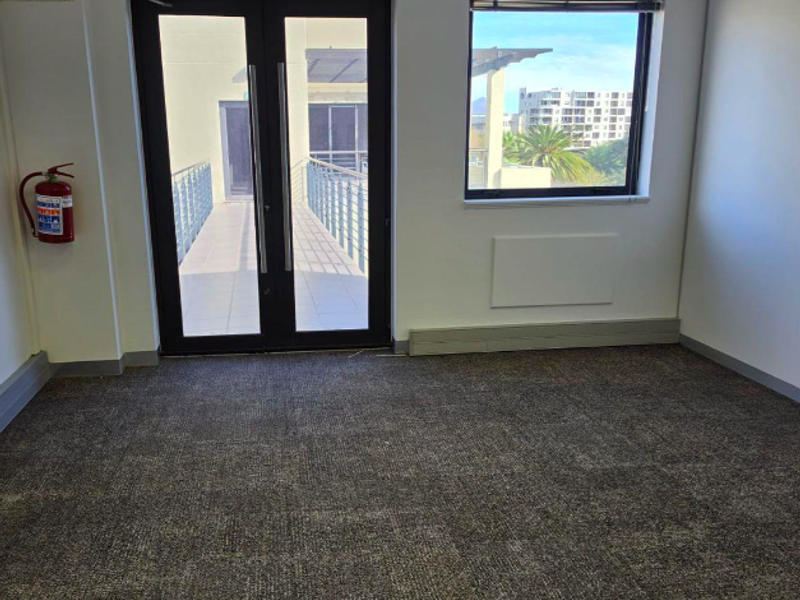 A clean, empty office space featuring carpet flooring, a wall-mounted heater, and double glass doors leading to an outdoor balcony area.