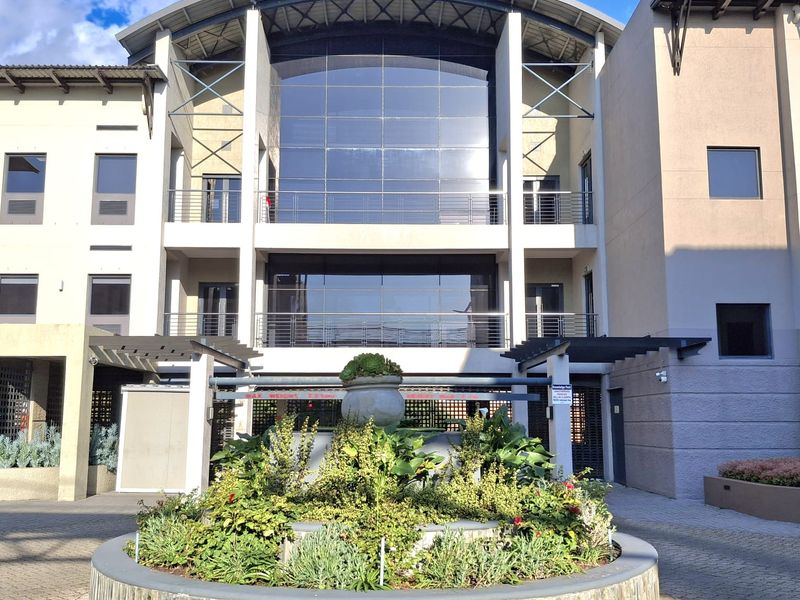 Modern office building facade featuring a prominent glass curtain wall, steel balcony railings, and a central stone-clad planter in the paved entrance courtyard.