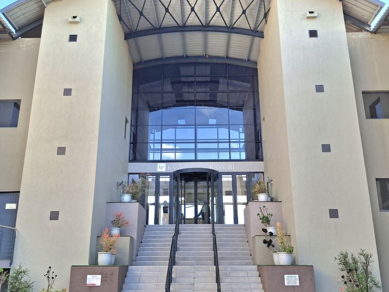 Front elevation of a modern office building featuring a prominent glass entrance, revolving doors, and a grand concrete staircase with metal handrails.