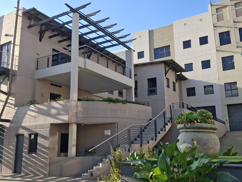 Exterior view of a modern office building featuring a raised balcony with a pergola, external staircase, and paved courtyard area.