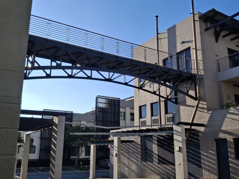 Exterior view of the office complex featuring a modern metal pedestrian bridge connecting building sections above a paved courtyard area.