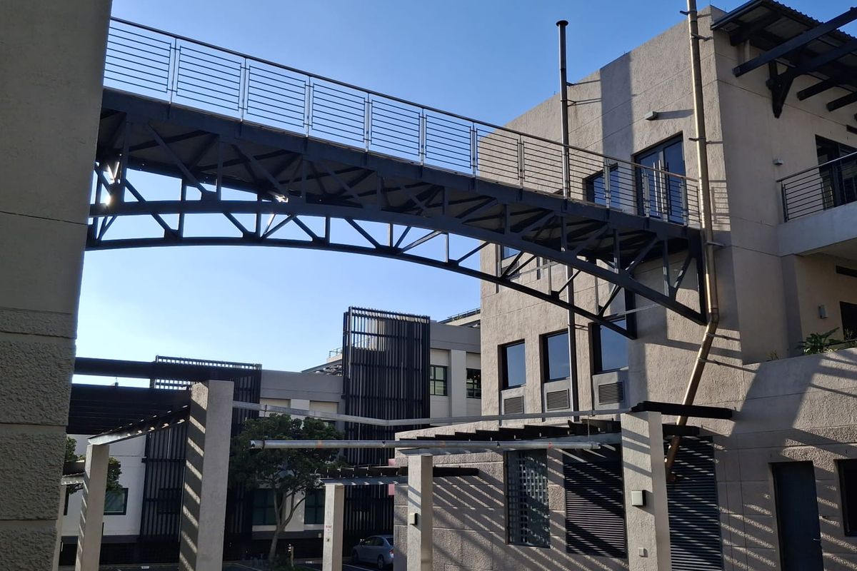 Exterior view of the office complex featuring a modern metal pedestrian bridge connecting building sections above a paved courtyard area.