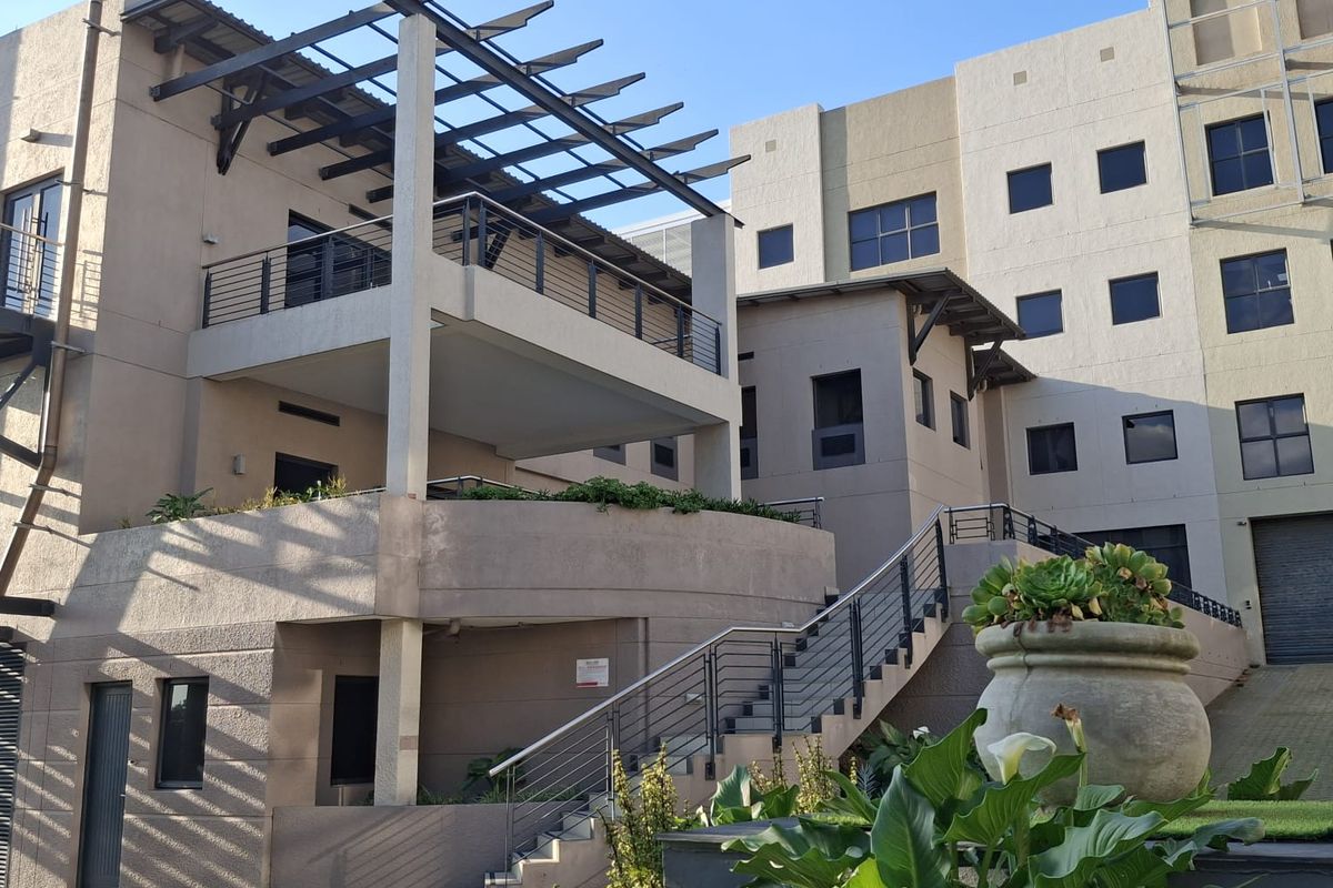 Exterior view of a modern office building featuring a raised balcony with a pergola, external staircase, and paved courtyard area.