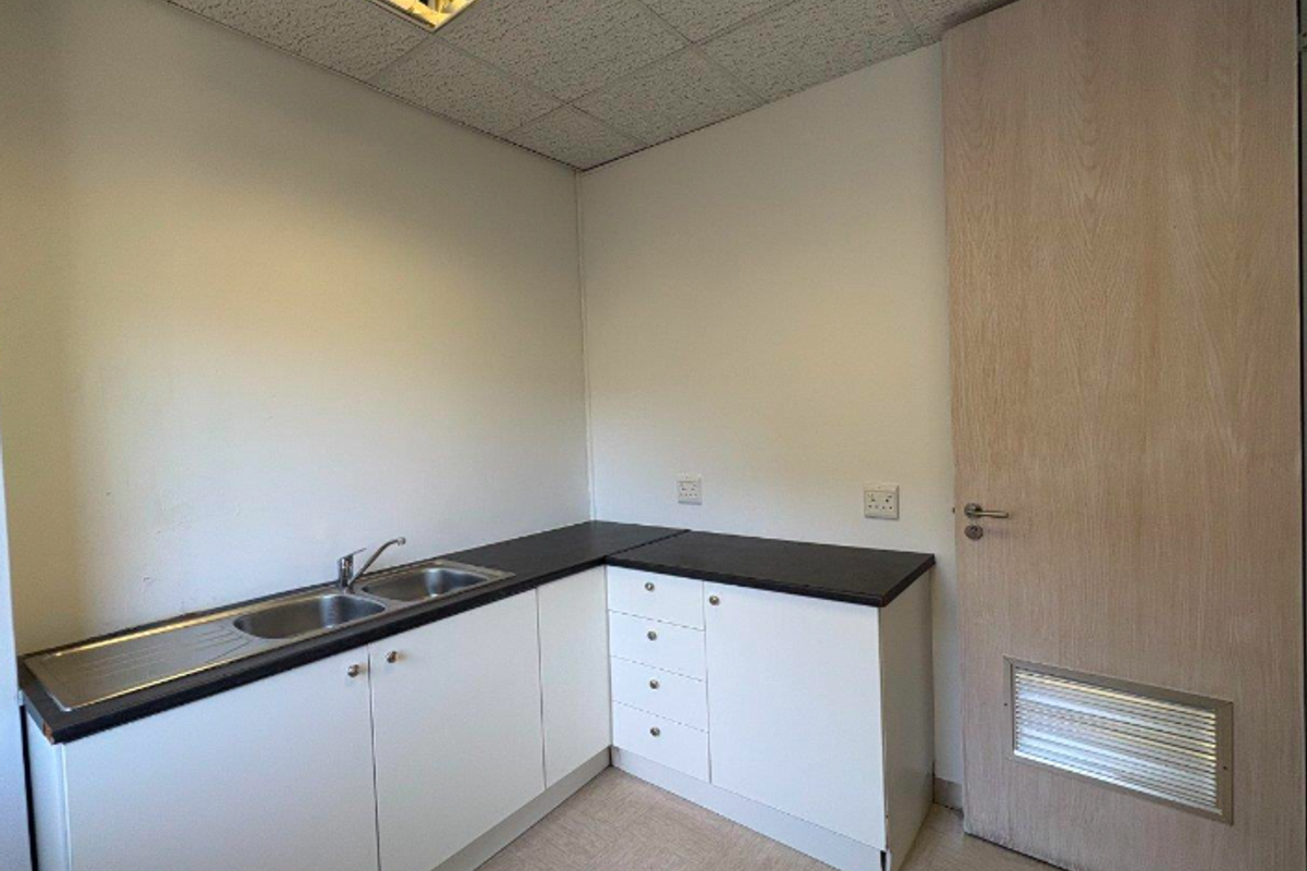 A functional office kitchenette featuring white cabinetry, dark laminate countertops, a stainless steel sink, and a drop ceiling with integrated lighting.