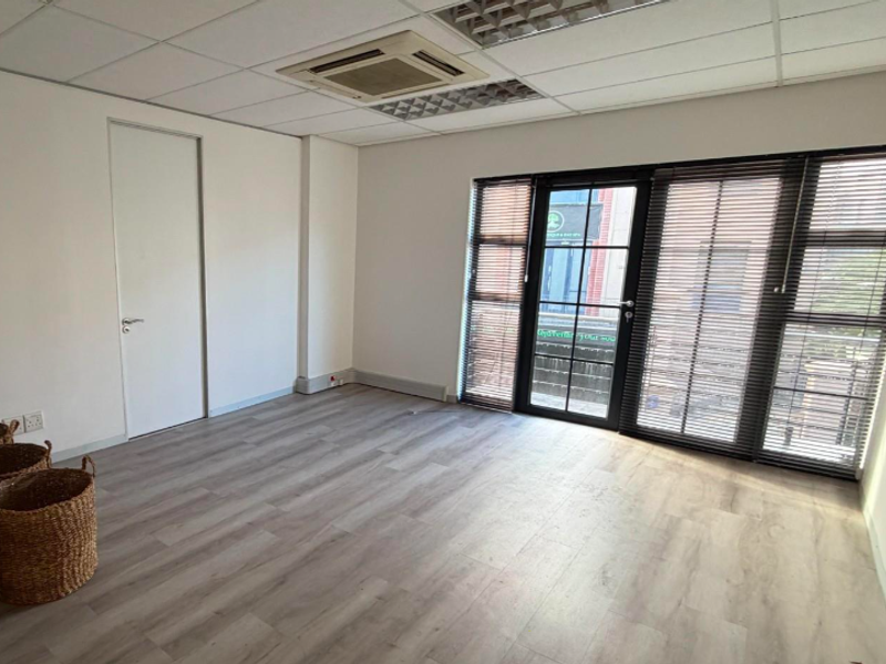 A bright, empty office space featuring modern laminate flooring, a suspended ceiling with integrated air conditioning, and large glass doors with venetian blinds.