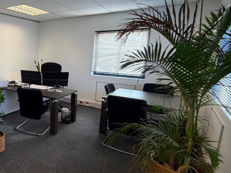 A functional office space featuring carpeted flooring, a suspended ceiling with recessed lighting, and windows fitted with venetian blinds.