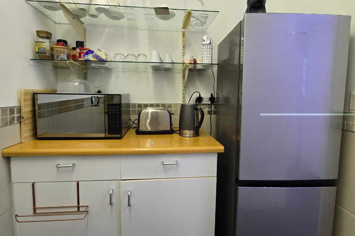 A compact office kitchenette featuring white cabinetry, a laminate countertop, tiled backsplash, and open glass shelving for storage.