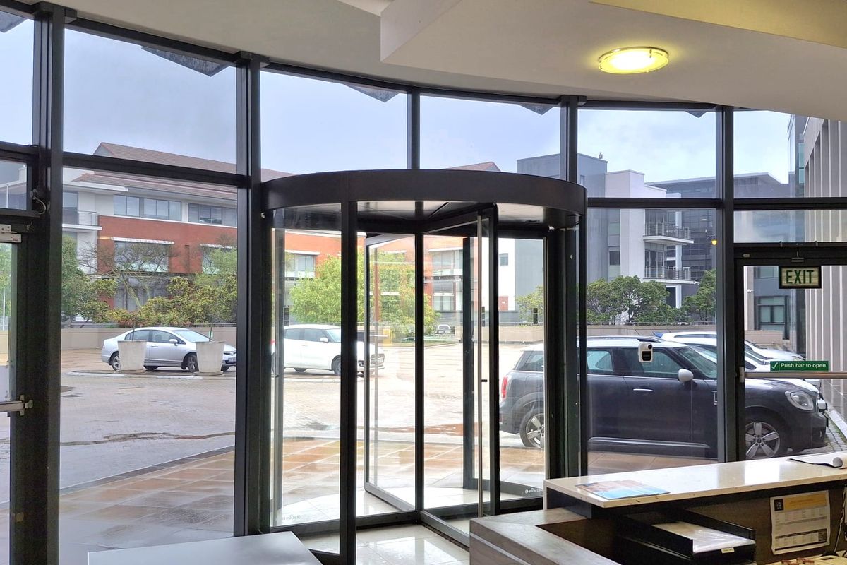 Modern office building entrance featuring a revolving door, floor-to-ceiling glass facade, security turnstile, and a professional reception desk area.