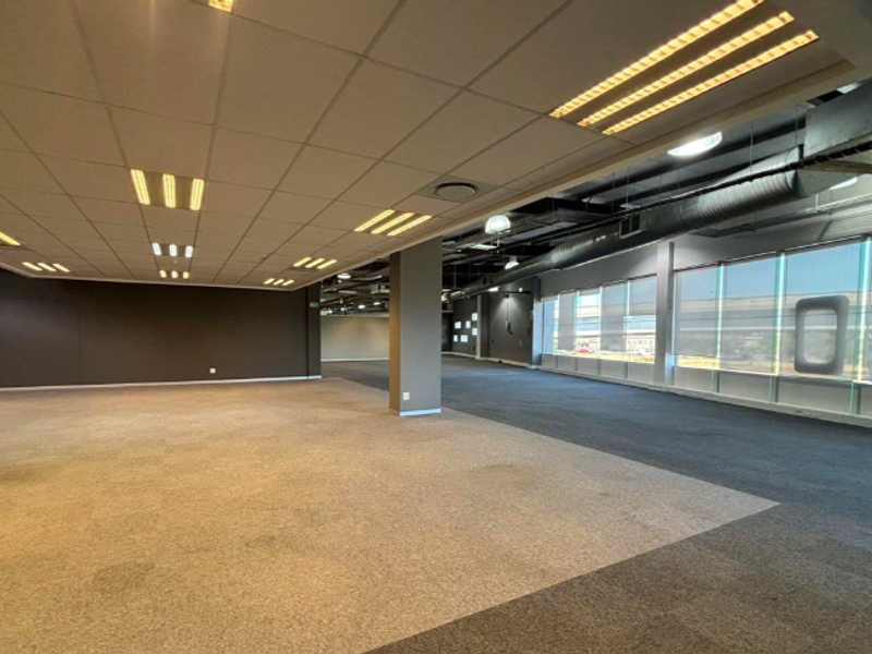 Spacious open-plan office area featuring suspended ceilings, exposed industrial ducting, large windows providing natural light, and durable carpet tile flooring.