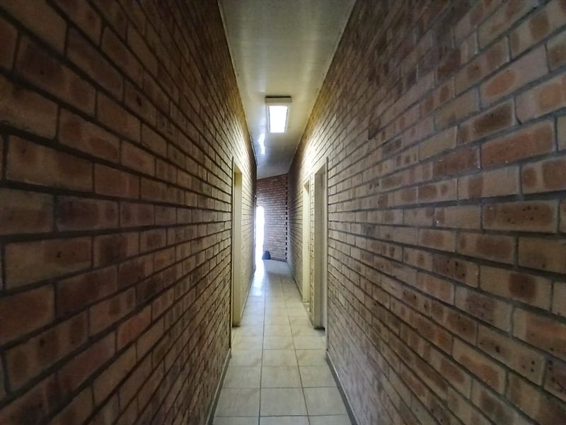 A long, narrow commercial hallway featuring exposed brick walls, ceramic tiled flooring, and a central fluorescent light fixture.