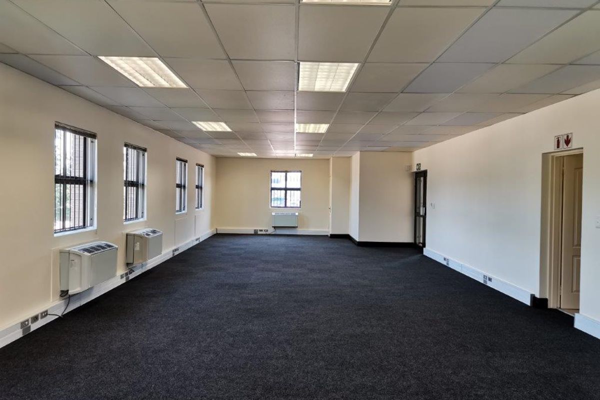 A spacious, open-plan office area featuring a suspended ceiling with recessed lighting, dark carpeting, and multiple wall-mounted air conditioning units.