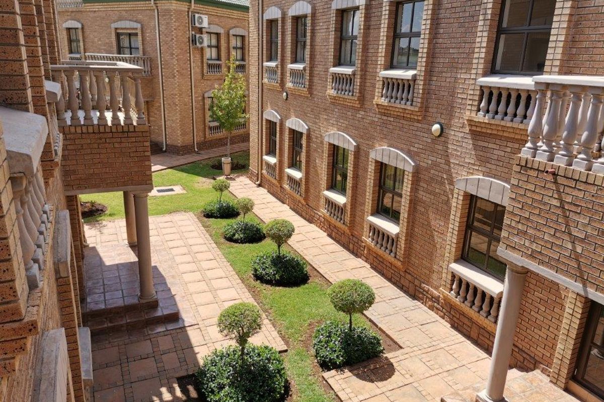 A well-maintained exterior courtyard featuring brick facade architecture, paved walkways, manicured garden beds, and classic balustrade detailing between office building wings.