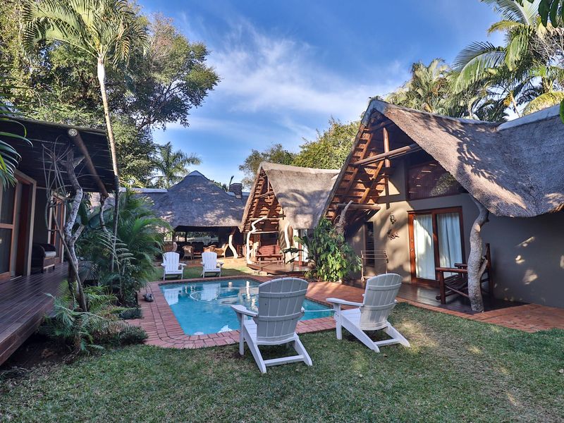 A tranquil outdoor pool area featuring thatched-roof structures, timber decking, and lush garden surroundings, perfect for relaxation in a guest house setting.