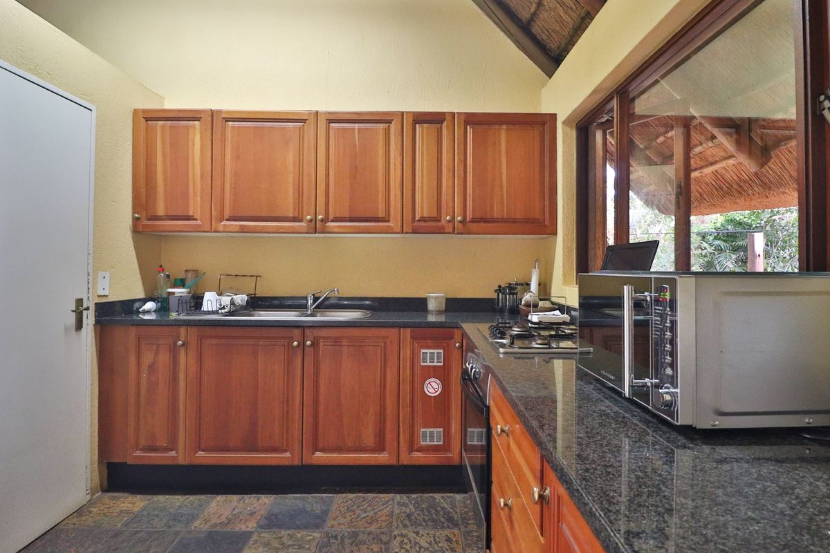 A functional kitchen featuring wooden cabinetry, granite countertops, a gas hob, and built-in oven, set under a traditional thatched roof.