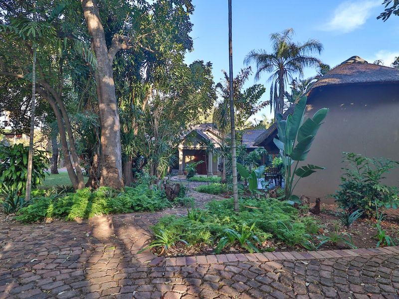 A lush, landscaped garden featuring a brick-paved pathway leading through mature trees toward a guest house structure with a traditional thatched roof.
