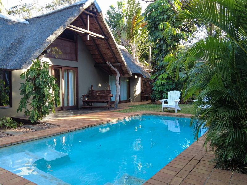 A well-maintained outdoor pool area featuring a swimming pool with brick coping, a wooden deck, and a thatched-roof structure with sliding glass doors.