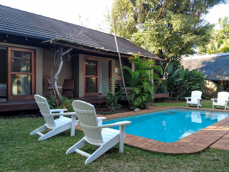 A well-maintained outdoor pool area featuring a swimming pool with brick coping, adjacent wooden decking, and access to the guest house via sliding doors.