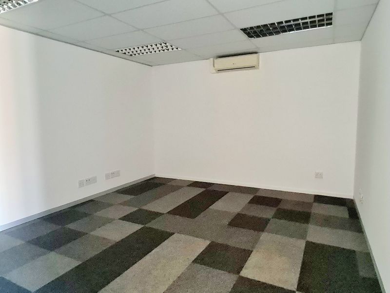 A clean, empty office space featuring carpet tile flooring, a suspended ceiling with recessed lighting, and a wall-mounted air conditioning unit.