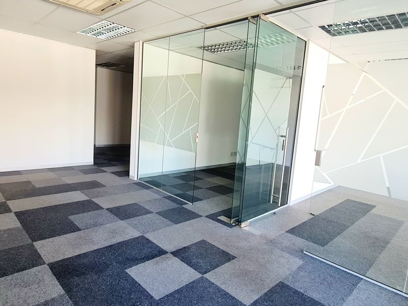 Modern open-plan office space featuring glass-partitioned meeting rooms, suspended ceilings with recessed lighting, and patterned carpet tiles.