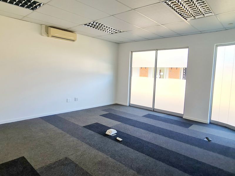 A bright, empty office space featuring carpet tiles, a suspended ceiling with recessed lighting, a split-system air conditioner, and a curved glass wall.