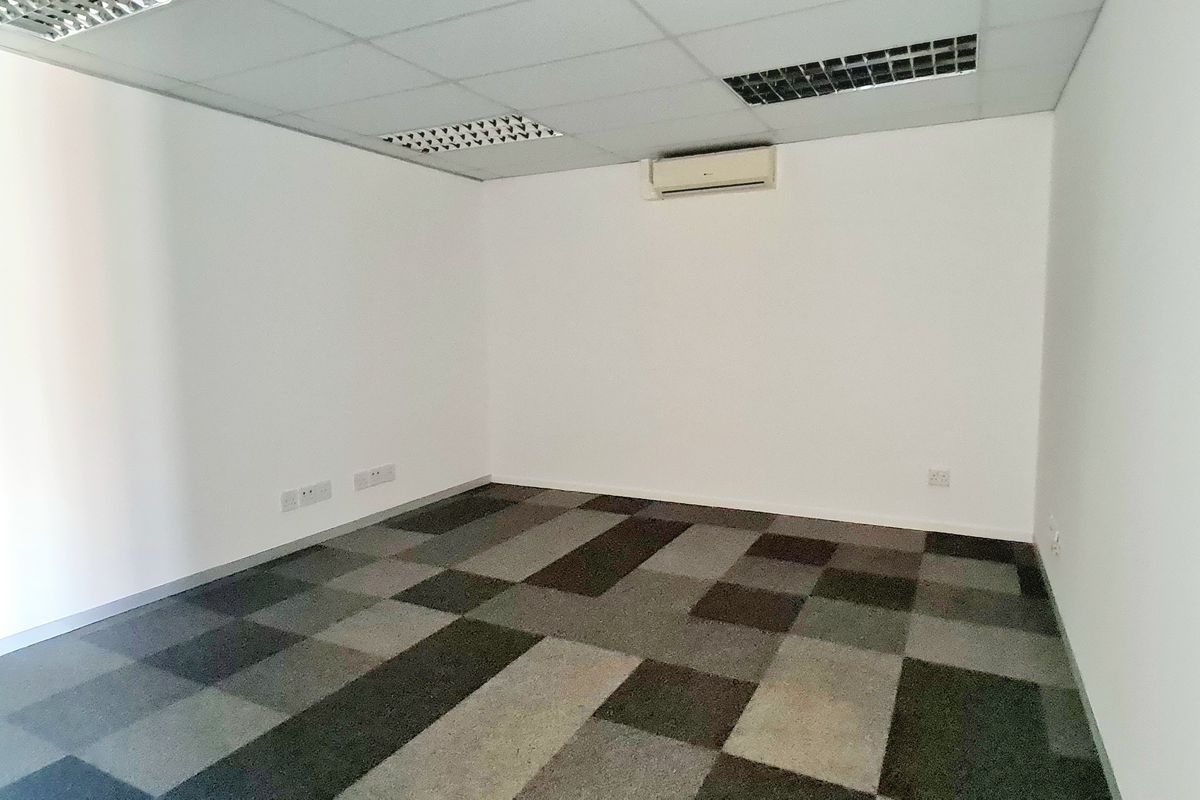 A clean, empty office space featuring carpet tile flooring, a suspended ceiling with recessed lighting, and a wall-mounted air conditioning unit.