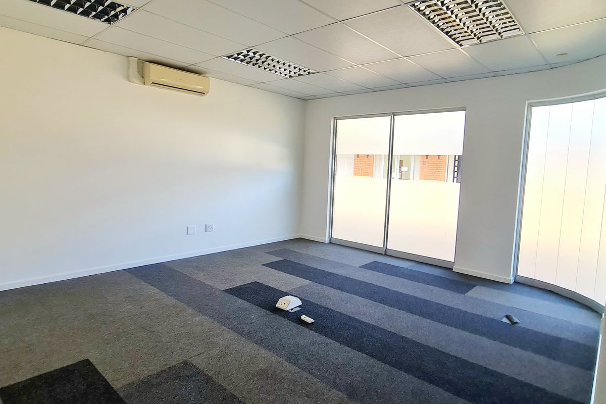 A bright, empty office space featuring carpet tiles, a suspended ceiling with recessed lighting, a split-system air conditioner, and a curved glass wall.