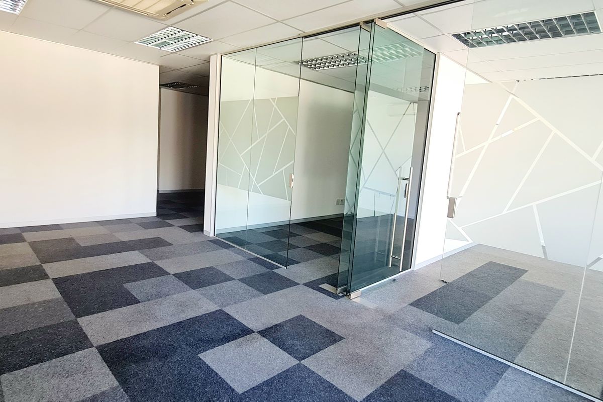 Modern open-plan office space featuring glass-partitioned meeting rooms, suspended ceilings with recessed lighting, and patterned carpet tiles.