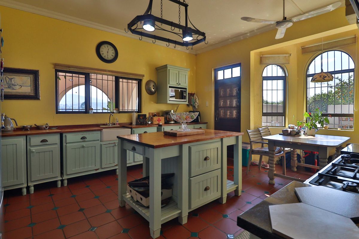 Spacious farmhouse kitchen featuring light green cabinetry, wooden countertops, a central island, farmhouse sink, and terracotta tile flooring with abundant natural light.