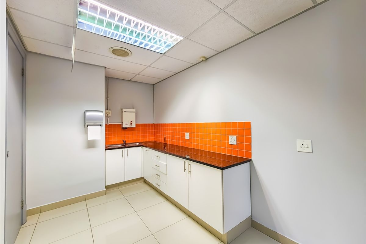 A functional office kitchenette featuring white cabinetry, dark countertops, orange tiled backsplash, and a drop ceiling with integrated lighting.