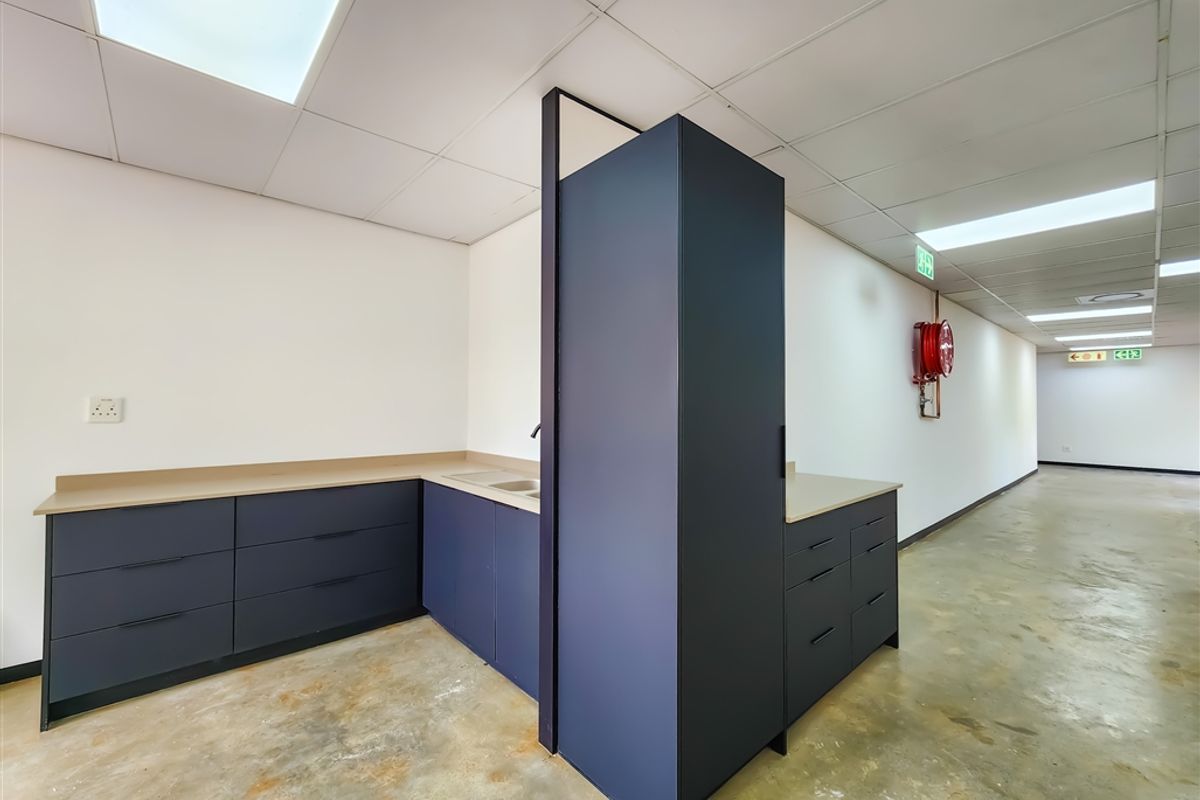 A functional office kitchenette area featuring modern dark blue cabinetry, integrated sink, and recessed ceiling lighting, situated adjacent to a main office corridor.