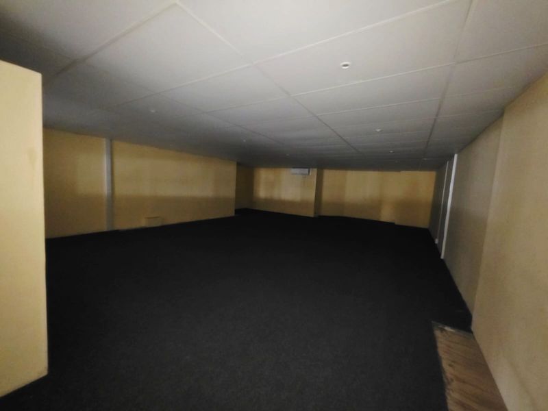 An empty, open-plan office space featuring a suspended ceiling, recessed lighting, split-system air conditioning, and dark carpet flooring.