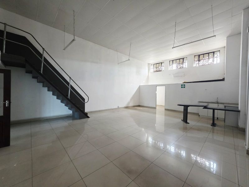 Spacious open-plan commercial area featuring tiled flooring, a metal staircase, a small kitchenette, and high-level windows providing natural light.