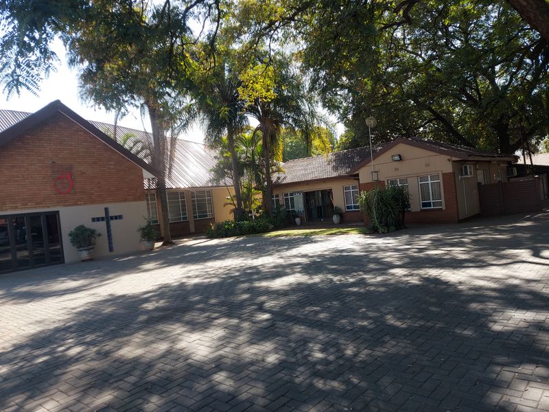 Front view of a commercial property featuring a brick facade, tiled roof, and a spacious paved driveway surrounded by mature trees.