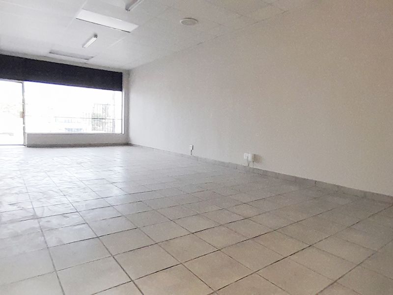 An empty, open-plan retail space featuring tiled flooring, a suspended ceiling with integrated lighting, and a large glass storefront window.